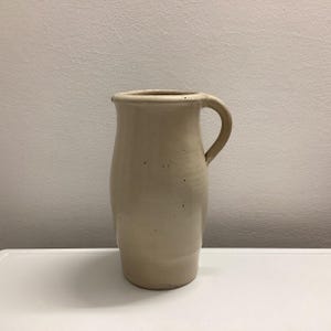 May include: A beige ceramic pitcher with a curved handle. The pitcher has a cylindrical body that tapers slightly towards the base. The opening is round, and the handle is attached near the top. The pitcher is sitting on a white surface.