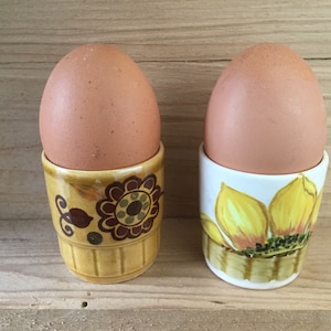 Könnte beinhalten: Zwei braune Eier in Keramik-Eierbechern. Ein Eierbecher hat ein gelbes und braunes Blumenmuster, der andere hat einen weißen Hintergrund mit einem gelben Sonnenblumenmuster.