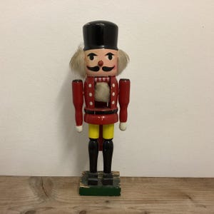 May include: A vintage wooden nutcracker with a red and yellow uniform, black hat, white beard, and a painted face. The nutcracker is standing on a green base.