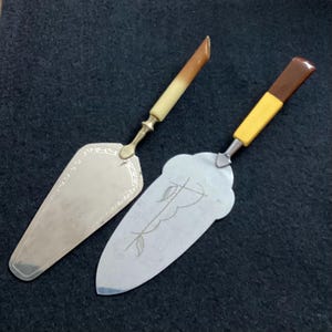 Könnte beinhalten: Zwei silberne Kuchenspachtel mit unterschiedlichen Griffen. Ein Griff ist braun und gelb, der andere ist cremefarben. Beide Spachtel haben ein dekoratives Design.
