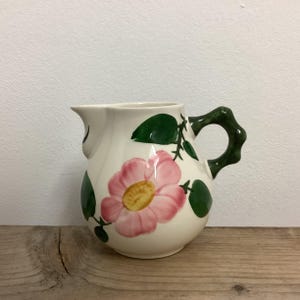 May include: A white ceramic jug with a green handle and a pink rose design. The jug features a floral pattern with green leaves and a single pink rose with a yellow centre.