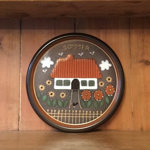 May include: A brown ceramic plate with a painted design of a white house with a red roof, surrounded by flowers and a fence. The plate has the word "SOMMER" written on it.
