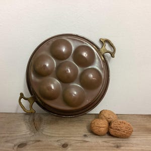May include: A copper escargot pan with seven round indentations and brass handles. The pan sits on a wooden surface with three walnuts.
