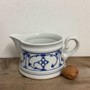 May include: White ceramic creamer with a blue floral design. The creamer has a spout and a handle. A walnut sits next to the creamer on a wooden surface. The creamer is a classic piece of kitchenware.