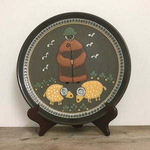May include: A decorative ceramic plate featuring a shepherd with a staff, two yellow sheep with white polka dots, and white birds. The plate has a dark brown rim and is displayed on a wooden stand.