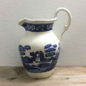 Könnte beinhalten: Eine weiße Keramikkanne mit einem blauen und weißen Blumendesign. Die Kanne hat eine breite Öffnung und einen Henkel, der sich nach oben und über die Oberseite krümmt. Das Design zeigt eine Landschaftsszene mit einer Brücke, Bäumen und einem Gebäude.