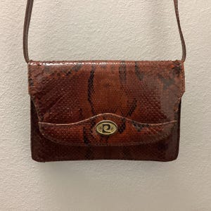 Könnte beinhalten: Eine braune Crossbody-Tasche aus Schlangenleder mit Klappenverschluss und goldenem Logo. Die Tasche hat einen langen, dünnen Riemen und eine rechteckige Form. Das Schlangenmuster weist dunkelbraune und schwarze Markierungen auf.