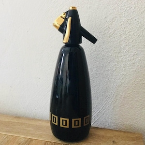May include: Black and gold ceramic soda siphon with a stylized bird head spout. The bottle has a geometric pattern of gold squares around the bottom.