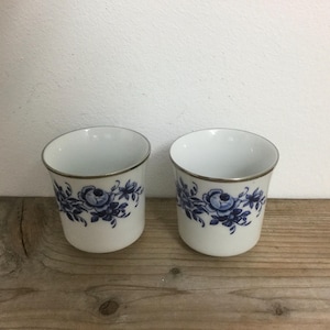 May include: Two white ceramic cups with a blue and silver floral design. The cups have a gold rim.