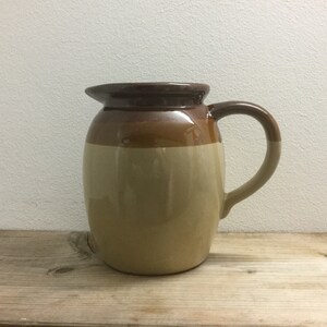 May include: A brown and beige ceramic jug with a rounded body and a handle. The jug has a glazed finish.