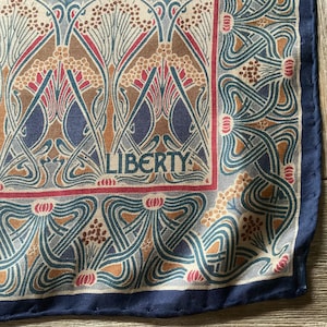May include: A square scarf featuring a repeating pattern of stylized floral motifs in shades of blue, tan, and cream, with a dark blue border. The word LIBERTY is printed in a dark blue serif font within the central design area of the fabric.