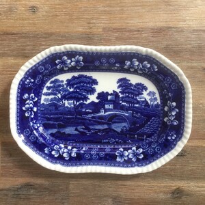 May include: An oval serving dish with a blue and white floral border and a blue and white landscape scene in the centre. The scene features a bridge, trees, and a building.