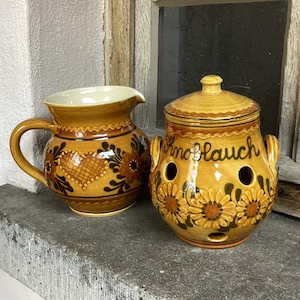 May include: A ceramic pitcher and garlic keeper set. The pitcher has a handle and floral designs. The garlic keeper has the word "Knoblauch" and sunflower designs. Both are in shades of brown and yellow.