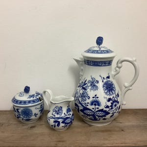 以下が含まれることがあります： 青い花柄の白い磁器のティーセット。 セットには、蓋付きのティーポット、クリーマー、蓋付きの砂糖入れが含まれます。 ティーポットには、湾曲したハンドルと注ぎ口があります。