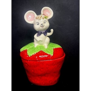 May include: A ceramic trinket box shaped like a strawberry, with a white mouse figurine on top. The mouse has pink-lined ears and a purple bow tie, sitting on green leaves. The box is a bright red colour.