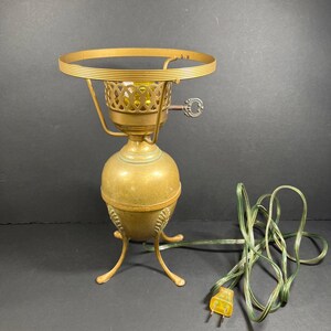 Op de afbeelding: Een vintage messing tafellamp met een decoratieve opengewerkte lampenkaphouder en een sleutelvormige schakelaar. De lamp heeft een eivormige voet en drie gebogen poten. Een groen elektrisch snoer met een gele stekker is bevestigd.
