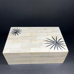 Puede incluir: Una caja rectangular crema con un patrón similar a ladrillos. La tapa presenta dos diseños de estrellas negras. Esta caja probablemente se utiliza para guardar o exhibir objetos pequeños.