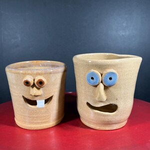 Puede incluir: Dos tazas de cerámica color beige con caras expresivas. Una taza tiene ojos marrones y un diente, la otra tiene ojos azules. Ambas tazas tienen bocas abiertas. Las tazas están sobre una superficie roja.