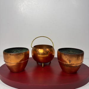 May include: A set of three copper vessels consisting of two open bowls with a dark green interior patina and one central spherical pot featuring a thin brass handle and three small feet, all displaying a polished metallic exterior finish.