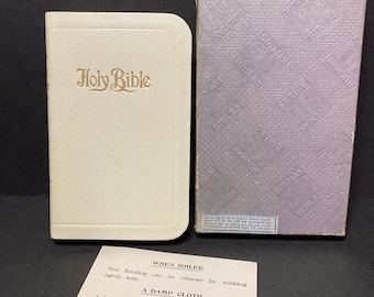 May include: A cream-colored Holy Bible with gold lettering, next to a matching box. The box is lavender with embossed text. A small paper with cleaning instructions is in front of the Bible.