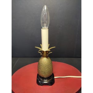 Puede incluir: Una lámpara de mesa con forma de piña de latón con un accesorio de luz tipo vela de color crema y una bombilla transparente. La lámpara se asienta sobre una base negra y tiene un cuerpo de piña dorado. La lámpara está enchufada a una toma de corriente.