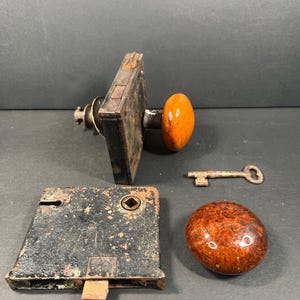 Op de afbeelding: Een antieke deurslotset met een roestige, rechthoekige metalen plaat, een bijpassende deurknop en een sleutel. De knop heeft een warme, oranjebruine kleur. De set bevat een tweede, kleinere knop en een sleutel.