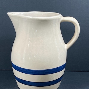 Op de afbeelding: Crèmekleurige keramische kan met twee horizontale blauwe strepen. De kan heeft een handvat en een schenktuit. Het ontwerp is eenvoudig en klassiek, geschikt voor het serveren van dranken of als decoratief item.