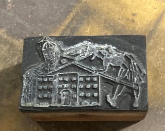 May include: A rectangular, vintage letterpress printing block. The block features a detailed relief of a building with multiple windows and a figure on horseback. The block is made of a dark material and mounted on a wooden base.
