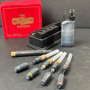 May include: A set of Koh-I-Noor Rapidograph technical pens, ink bottle, and accessories. The pens have clear barrels, gray bodies, and gold accents. The set includes a red box with gold lettering and a black pen holder.