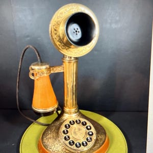 以下が含まれることがあります： 真鍮製のアンティーク燭台電話、ロータリーダイヤル付き。 電話は、ベースと受話器に装飾的なデザインが施され、黒いイヤホンとオレンジ色のマウスピースが付いています。 電話は緑色の円形ベースに置かれています。