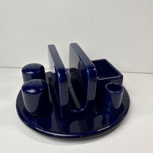 Puede incluir: Un juego de comedor de cerámica azul marino con acabado brillante. El juego incluye un salero, un pimentero, un servilletero, un pequeño recipiente rectangular y un pequeño recipiente cilíndrico, todo dispuesto sobre una base circular.