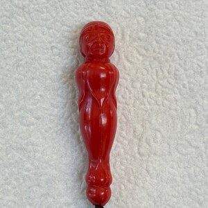 May include: A bright red, elongated, carved object featuring a stylized human-like figure with a rounded head, defined facial features, and a smooth, tapered body that ends in a small, spherical base, crafted from a solid, glossy material.