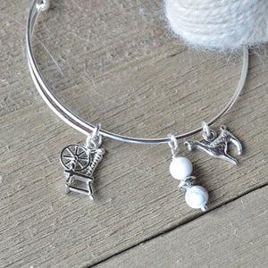 Könnte beinhalten: Ein silbernes Armreif-Armband mit drei Anhängern: einem Spinnrad, zwei weißen Perlen und einem kleinen silbernen Reh. Das Armband liegt auf einer Holzoberfläche.