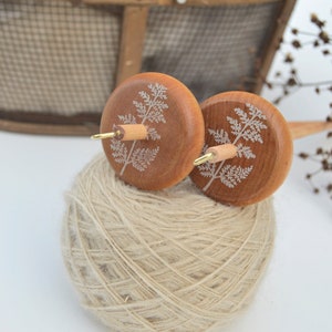 May include: Two wooden spinning wheels with a fern design on each wheel. The wheels are attached to a wooden spindle. The spindle is resting on a ball of beige yarn.