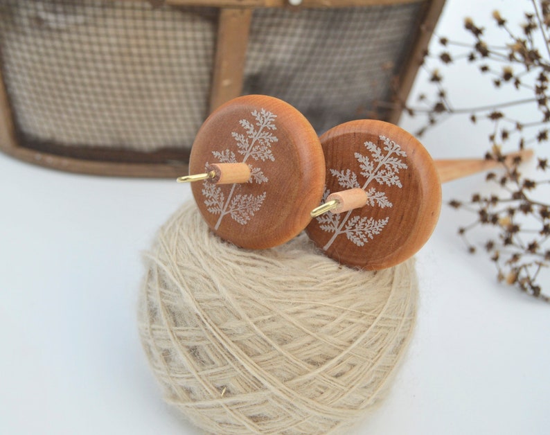 May include: Two wooden spinning wheels with a fern design on each wheel. The wheels are attached to a wooden spindle. The spindle is resting on a ball of beige yarn.