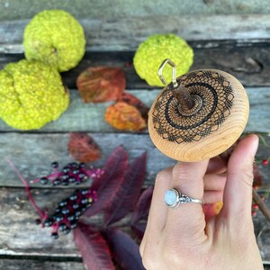 May include: A wooden spinning wheel with a black and white geometric design. The wheel is held in a hand wearing a silver ring with a white stone.