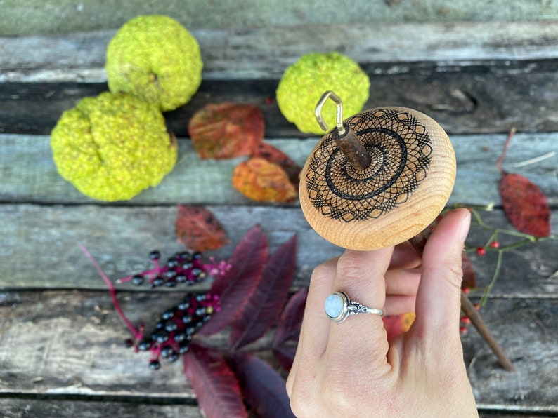 May include: A wooden spinning wheel with a black and white geometric design. The wheel is held in a hand wearing a silver ring with a white stone.