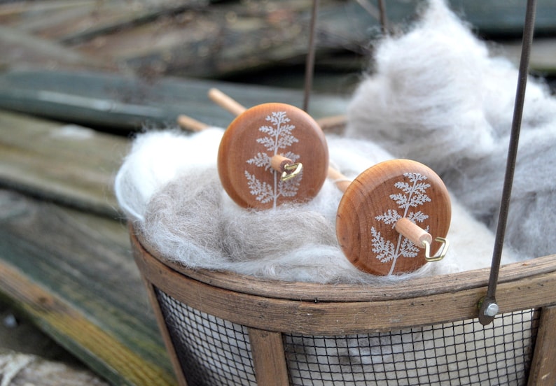 May include: Two wooden spinning wheels with a fern design on the top. The wheels are in a basket filled with white fluffy fiber.