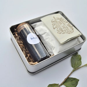 May include: A silver tin box filled with natural dye supplies. The box contains a small glass jar labeled "Indigo", a white powder, and a folded card with a brown pine cone illustration.