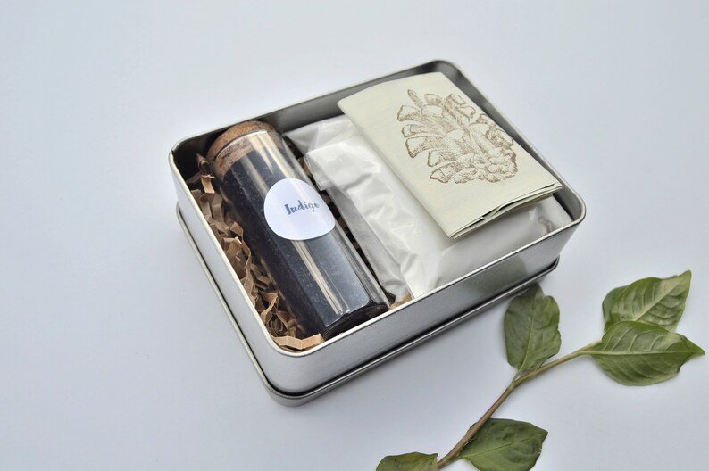 May include: A silver tin box filled with natural dye supplies. The box contains a small glass jar labeled "Indigo", a white powder, and a folded card with a brown pine cone illustration.