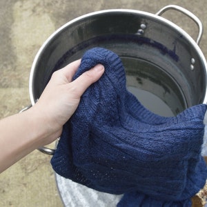 May include: A hand holding a dark blue knitted sweater over a stainless steel pot filled with water.