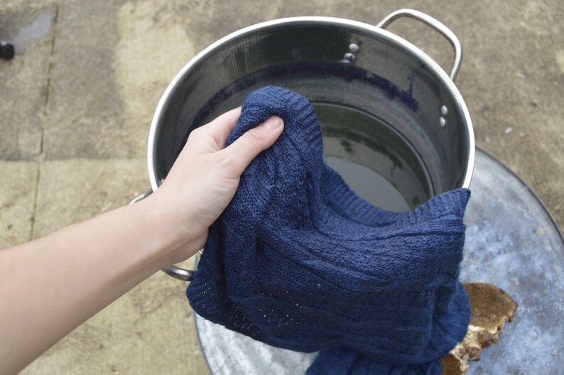 May include: A hand holding a dark blue knitted sweater over a stainless steel pot filled with water.