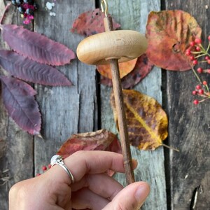 May include: A wooden spinning wheel with a natural wood finish and a gold metal hook. The wheel is held in a hand against a background of wooden planks and autumn leaves.