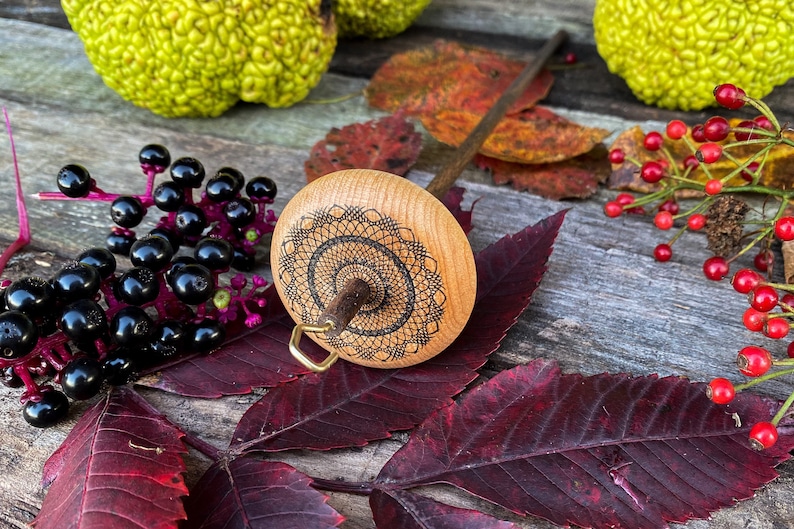 May include: A wooden spinning wheel with a black and white mandala design on the whorl. The wheel is resting on a wooden surface with fall leaves and berries.