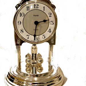 May include: A silver-toned Kundo quartz clock with a glass dome and a pendulum. The clock face has black Roman numeral hour markers and black hands.
