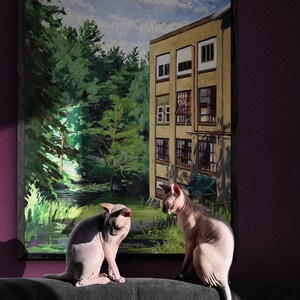May include: Two hairless cats sit on a dark gray couch in front of a framed painting of a building with a green forest behind it. The painting has a blue sky with white clouds.