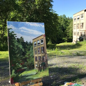 May include: An easel with a painting of a building with trees in the foreground. The painting is set up outdoors in front of a similar building with trees in the background.
