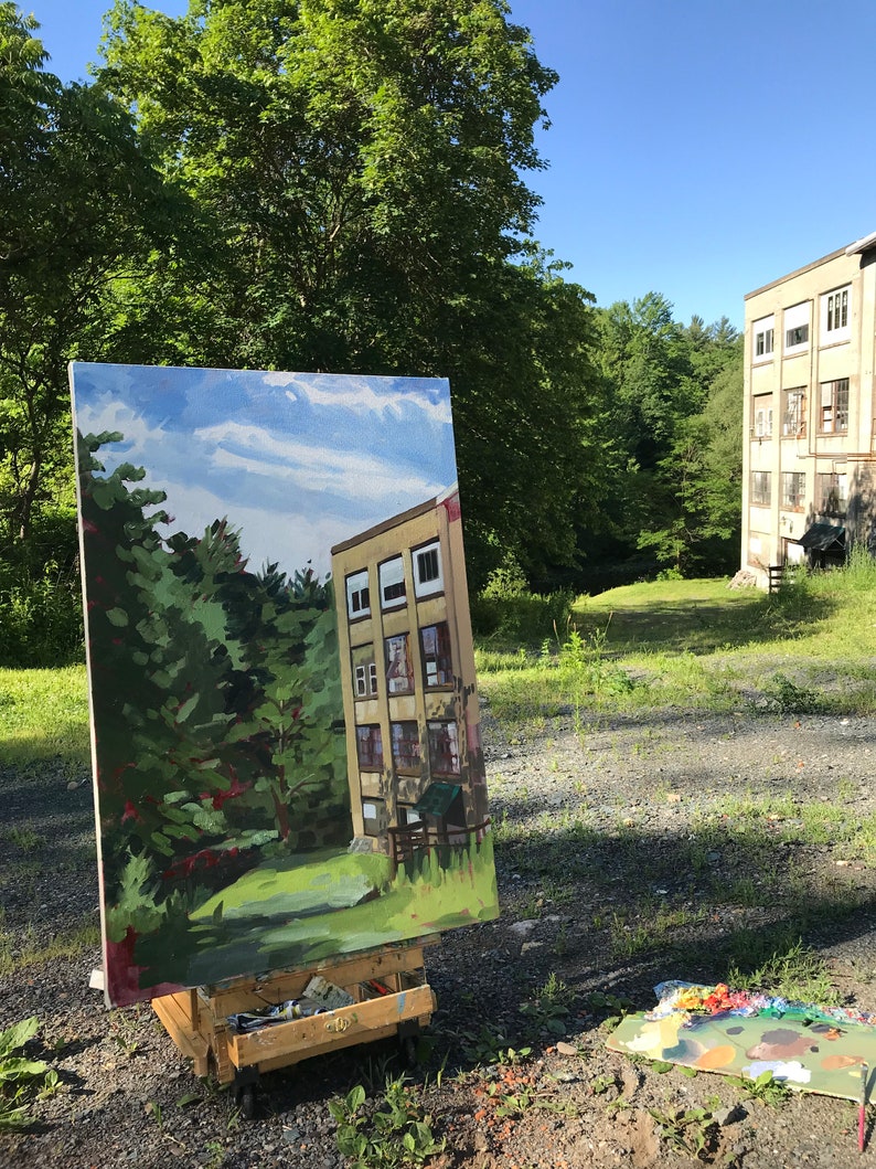 May include: An easel with a painting of a building with trees in the foreground. The painting is set up outdoors in front of a similar building with trees in the background.