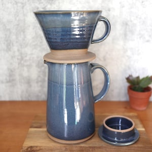 May include: A handmade ceramic pour-over coffee set in shades of blue. The set includes a dripper, a carafe with a handle, and a lid. The pieces are glazed with a glossy finish and sit on a wooden surface. A small succulent plant is in the background.