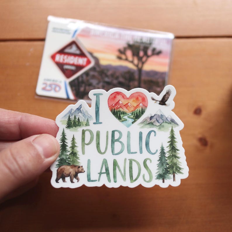 May include: A white sticker with the words "I Public Lands" and a heart-shaped graphic of mountains, trees, and a river. A brown bear and an eagle are also featured. A card with the word "Resident" is in the background.